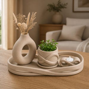 Könnte beinhalten: Ein beiges Keramikset bestehend aus einem ovalen Tablett mit geschlungenem Rand, einer ringförmigen Vase mit getrockneten Gräsern, einem kleinen Pflanzgefäß mit einer grünen Sukkulente und einem passenden Kerzenhalter mit weißem Teelicht.