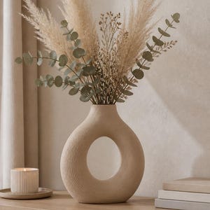 Könnte beinhalten: Eine beige, ringförmige Vase mit einer texturierten, gerippten Oberfläche und einer zentralen kreisförmigen Öffnung. Die Vase ist mit einem Strauß aus getrocknetem Pampasgras und grünen Eukalyptuszweigen gefüllt, was ein luftiges Arrangement ergibt.