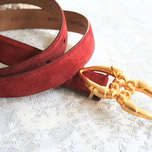 May include: A red suede belt with a gold-colored buckle. The belt is medium-sized and made of genuine leather. The buckle has an ornate design.