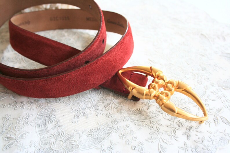 May include: A red suede belt with a gold-colored buckle. The belt is medium-sized and made of genuine leather. The buckle has an ornate design.
