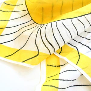 May include: A square scarf featuring a white background with bold yellow borders and black hand-drawn style wavy lines radiating outward. The fabric has a semi-sheer appearance and includes the signature text Varia in the bottom left corner.
