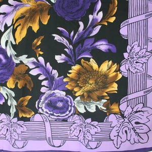 Puede incluir: Un pañuelo cuadrado con un estampado floral. El diseño presenta flores y hojas moradas, doradas y blancas sobre un fondo negro. El borde es lavanda con un diseño de cinta y hojas.