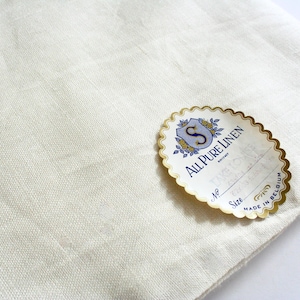 May include: Off-white linen fabric with a textured weave. An oval label with gold trim and the text "All Pure Linen" is on the fabric. The label also includes the text "Made in Belgium".
