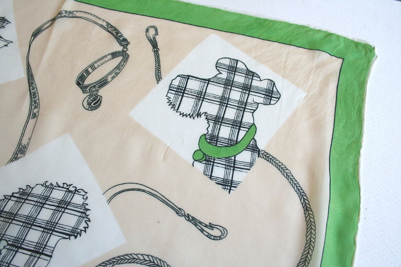 May include: A cream-colored fabric with a green border. The fabric features a black and white plaid design of a dog, a collar, and a leash.