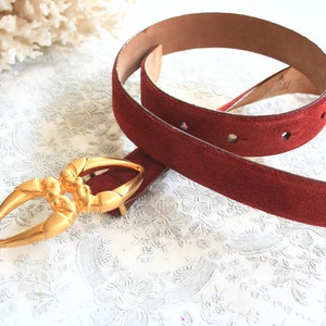 May include: A burgundy suede belt with a gold-toned buckle. The belt has a textured surface and several holes for adjustment. The buckle is an ornate design. The belt is displayed on a white floral patterned surface.
