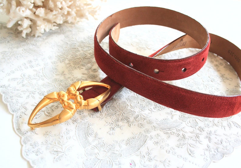 May include: A burgundy suede belt with a gold-toned buckle. The belt has a textured surface and several holes for adjustment. The buckle is an ornate design. The belt is displayed on a white floral patterned surface.