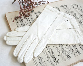 May include: A pair of off-white leather gloves featuring decorative stitching along the back of the hand and a small, subtle floral-like detail near the wrist. The gloves have a smooth texture and are positioned overlapping each other.