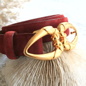May include: A burgundy suede belt with a gold-colored buckle. The buckle has an ornate design. The belt is rolled up, showcasing the texture of the suede. The background is a light color.