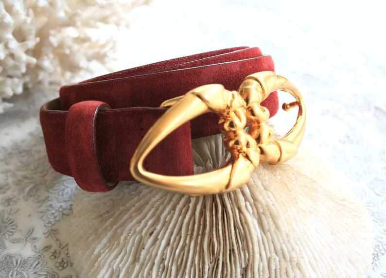 May include: A burgundy suede belt with a gold-colored buckle. The buckle has an ornate design. The belt is rolled up, showcasing the texture of the suede. The background is a light color.