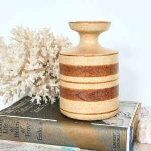 May include: A beige ceramic vase with horizontal brown stripes. The vase has a flared top and a cylindrical body. It is sitting on a vintage book with gold lettering and a white coral decoration.