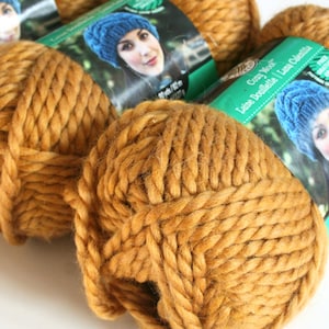 May include: Close-up of several skeins of chunky, golden-brown yarn. Each skein is tightly wound, showing the thick texture of the wool. Green and white labels with a woman wearing a blue hat are visible on the yarn.
