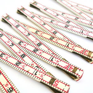 May include: Several vintage folding rulers are arranged diagonally against a white background. The rulers are white with red markings and black numbers, measuring up to 9 inches. The rulers are made of wood and have metal joints.