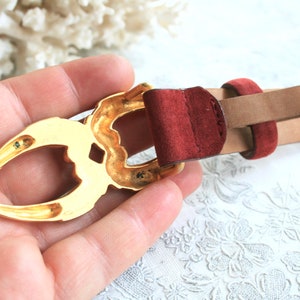 May include: A tan leather belt with a gold-colored buckle. The buckle has an ornate design and is paired with a burgundy suede loop. The belt is a classic accessory.