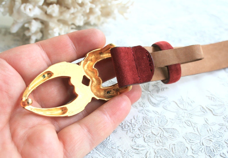 May include: A tan leather belt with a gold-colored buckle. The buckle has an ornate design and is paired with a burgundy suede loop. The belt is a classic accessory.