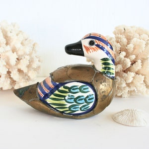 May include: A decorative duck figurine with a brass body and a ceramic head. The head features a black beak, and colorful stripes in blue, orange, and green. The duck is displayed with white coral and a seashell.