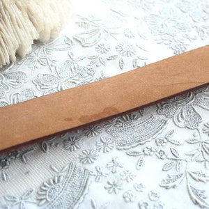 May include: A close-up of a light brown leather belt. The belt is rectangular and has a slightly textured surface. The background is a white, floral-patterned fabric.