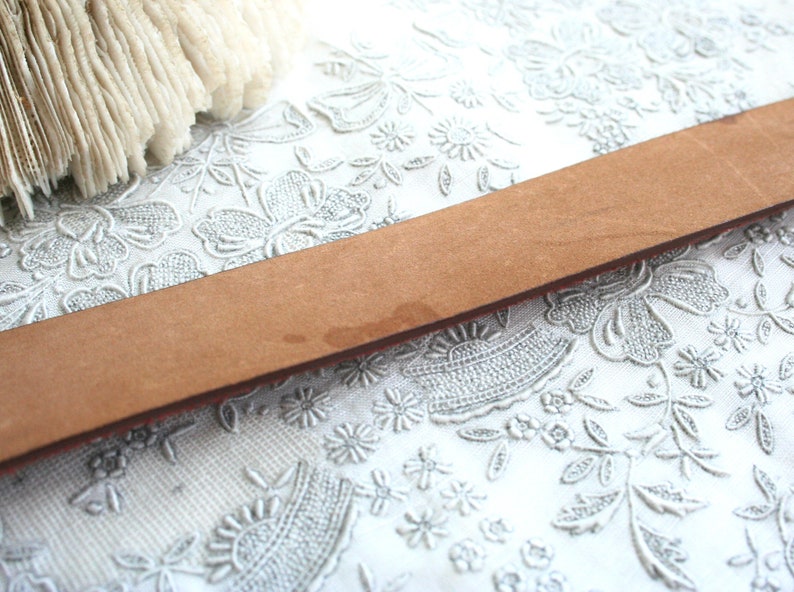 May include: A close-up of a light brown leather belt. The belt is rectangular and has a slightly textured surface. The background is a white, floral-patterned fabric.