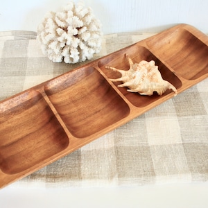 May include: A rectangular wooden serving tray with four compartments. A seashell rests in one compartment. The tray is made of light brown wood and sits on a checkered beige and white cloth. A white coral decoration is also present.