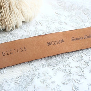 May include: Close-up of a medium-sized, brown leather belt. The belt is stamped with "MEDIUM" and "Genuine Leather." The code "G2C1035" is also visible. The belt is set against a white, floral-patterned background.
