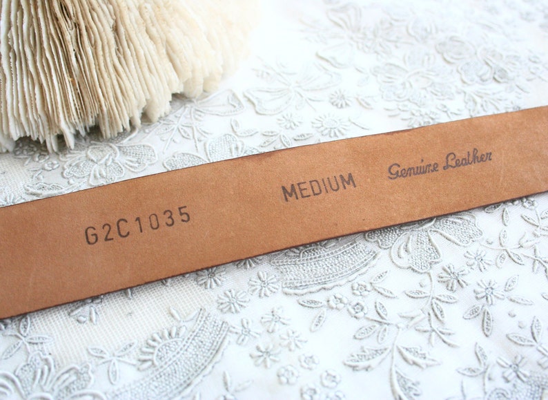 May include: Close-up of a medium-sized, brown leather belt. The belt is stamped with "MEDIUM" and "Genuine Leather." The code "G2C1035" is also visible. The belt is set against a white, floral-patterned background.