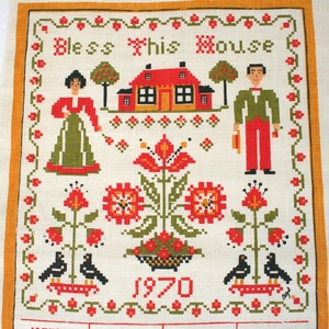 May include: Embroidered wall hanging with the text "Bless This House" and the year 1970. The design features a red house, figures in traditional clothing, flowers, and birds, all in a folk art style. The border is orange.