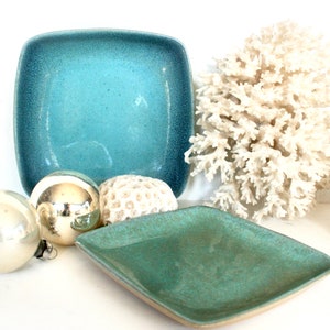 May include: Two square ceramic dishes in a teal green glaze. The dishes are sitting on a white surface with silver ornaments and a piece of white coral.