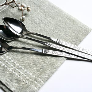 May include: Three silver spoons with ornate handle designs rest on a light green linen napkin. The spoons are arranged diagonally, reflecting light. A sprig of white berries adds a touch of natural detail to the composition.
