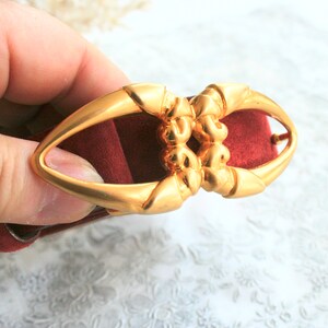 May include: A gold-colored metal belt buckle with an oval shape and ornate detailing. The buckle is held against a burgundy-colored fabric, showcasing its design and color contrast. The buckle's design is symmetrical and features a central decorative element.