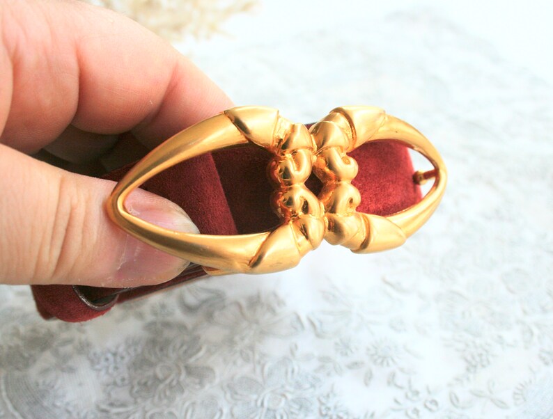 May include: A gold-colored metal belt buckle with an oval shape and ornate detailing. The buckle is held against a burgundy-colored fabric, showcasing its design and color contrast. The buckle's design is symmetrical and features a central decorative element.