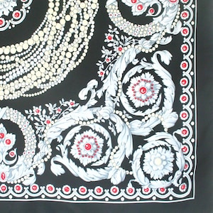Puede incluir: Pañuelo cuadrado negro con un patrón detallado. El diseño presenta motivos florales y de hojas blancos, hileras de perlas y detalles en rojo. Un borde blanco y rojo enmarca el pañuelo.