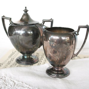 May include: A silver sugar and creamer set with ornate designs. The pieces have a polished finish and are sitting on a white tablecloth.