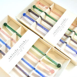May include: Boxes of swizzle sticks, each containing four sticks. The sticks have colorful glass ends in shades of pink, blue, green, and brown. The text "swizzle sticks set of 4" is printed on a white label.