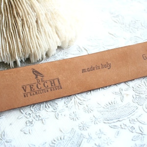 May include: A close-up of a brown leather belt with the text "made in Italy" and the brand "De Vecchi by Hamilton Hodge" printed on it. The belt has the code "G2C1035" printed on it. The background is a white floral pattern.