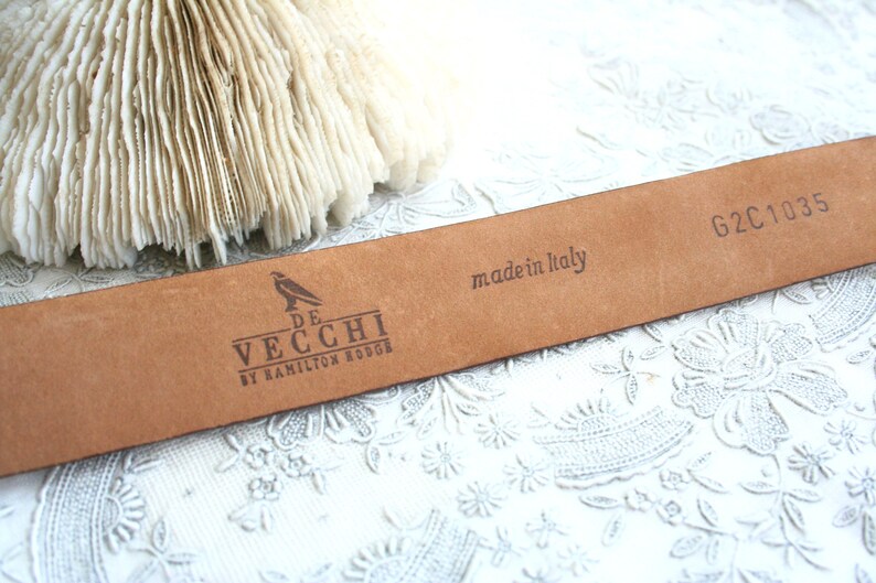 May include: A close-up of a brown leather belt with the text "made in Italy" and the brand "De Vecchi by Hamilton Hodge" printed on it. The belt has the code "G2C1035" printed on it. The background is a white floral pattern.