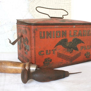 May include: An antique red metal container with the words "UNION LEADER CUT PLUG" and an eagle graphic. A wooden-handled tool and a metal tool with a wooden handle are in front of the container. The items are on a white surface.