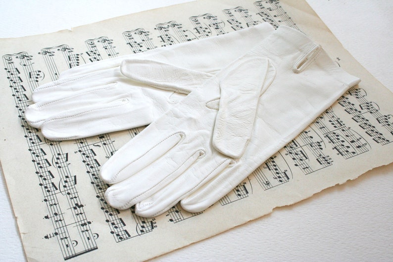May include: A pair of white leather gloves with a simple design. The gloves are laid out on a sheet of music.