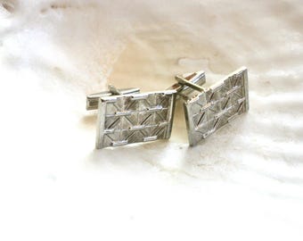 May include: A pair of silver-toned cufflinks with a rectangular shape and a textured, geometric design. The cufflinks are displayed on a white, textured surface, suggesting a vintage or classic style.