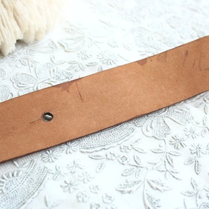 May include: A brown leather belt with two holes and some wear marks. The belt is a light brown color and appears to be vintage. The background is a white lace fabric.