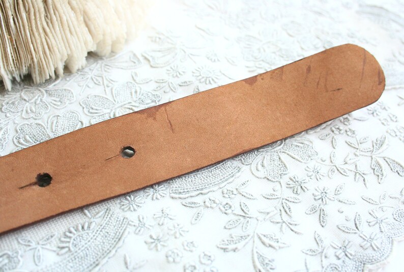 May include: A brown leather belt with two holes and some wear marks. The belt is a light brown color and appears to be vintage. The background is a white lace fabric.