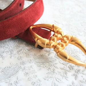 May include: A burgundy suede belt with a gold-colored buckle. The buckle has an ornate, open design. The belt is on a white, floral-patterned surface. The belt is a fashion accessory.