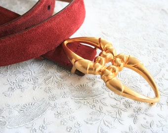 May include: A burgundy suede belt with a gold-colored buckle. The buckle has an ornate, open design. The belt is on a white, floral-patterned surface. The belt is a fashion accessory.