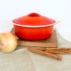 May include: A red enameled cooking pot with a lid, sitting on a stack of books. An onion and several cinnamon sticks are arranged nearby on a light gray patterned cloth. Kitchen and cooking theme.