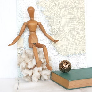 May include: A wooden artist's mannequin sits atop a white coral formation, with a vintage map of San Francisco in the background. A green book and a decorative metal sphere complete the still life arrangement.