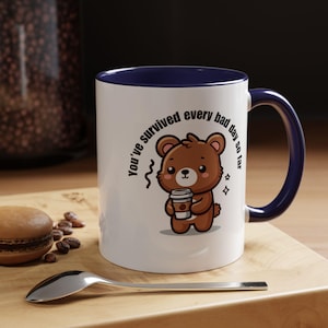 May include: White ceramic coffee mug with a blue rim and handle. The mug features a cartoon brown bear holding a cup of coffee and the text "You've survived every bad day so far".