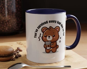 Op de afbeelding: Witte keramische koffiemok met een blauwe rand en handvat. De mok heeft een cartoonbruine beer die een kopje koffie vasthoudt en de tekst "You've survived every bad day so far".