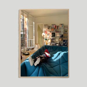 May include: A person is lying on a blue, quilted sofa in a room with a bookshelf and a window. The sofa is made of many small, quilted squares. The person is wearing a red shirt and jeans. The room is bright and sunny.