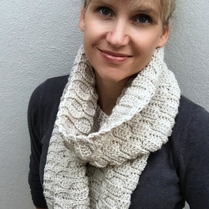 Peut inclure: Une femme portant un pull gris avec une écharpe en crochet blanc. L'écharpe a un motif texturé et est drapé autour de son cou.