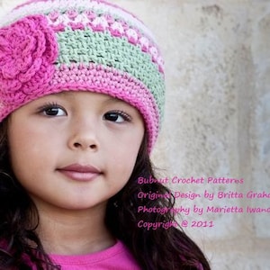 May include: A young girl wearing a pink, green, and white crocheted hat with a large pink flower on top. The hat has a striped pattern and is made with a soft yarn.