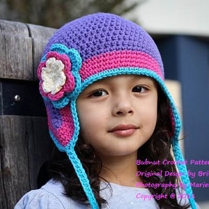 May include: A young person wearing a purple, pink, and blue crocheted hat with a flower embellishment. The hat has ear flaps and a long tie. The text "Bubnut Crochet Patterns Original Design by Britta Graham Photography by Marietta Iwanowski Copyright @ 2014" is visible at the bottom of the image.
