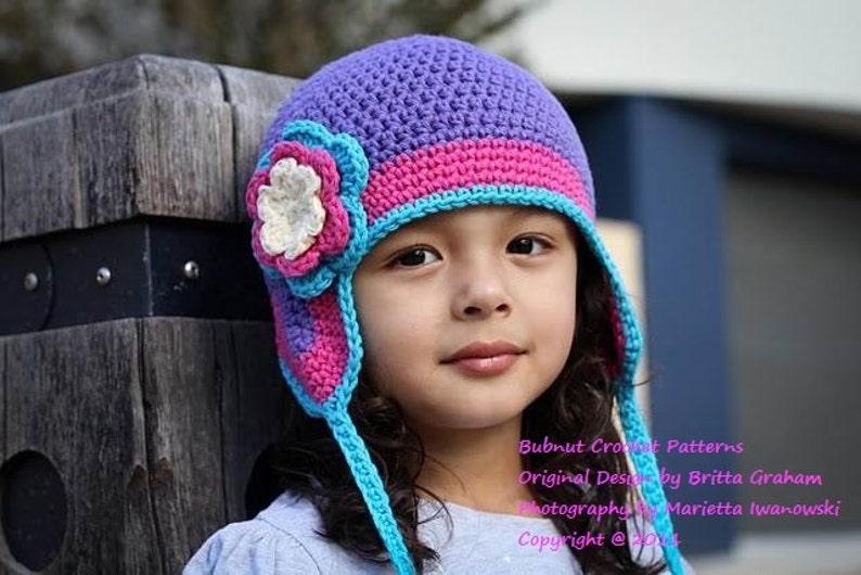 May include: A young person wearing a purple, pink, and blue crocheted hat with a flower embellishment. The hat has ear flaps and a long tie. The text "Bubnut Crochet Patterns Original Design by Britta Graham Photography by Marietta Iwanowski Copyright @ 2014" is visible at the bottom of the image.